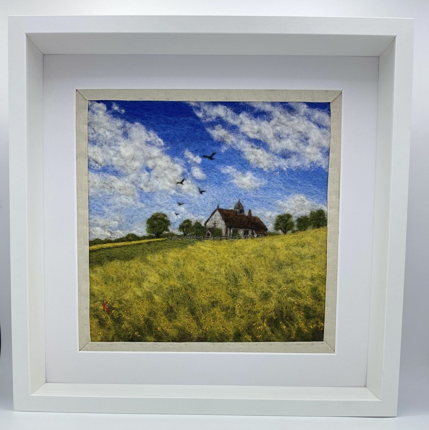 needle felted picture of a Medieval church in a field surrounded by yellow flowers with a blue sky above