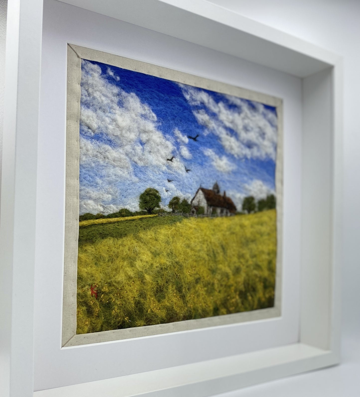 needle felted picture of a Medieval church in a field surrounded by yellow flowers with a blue sky above