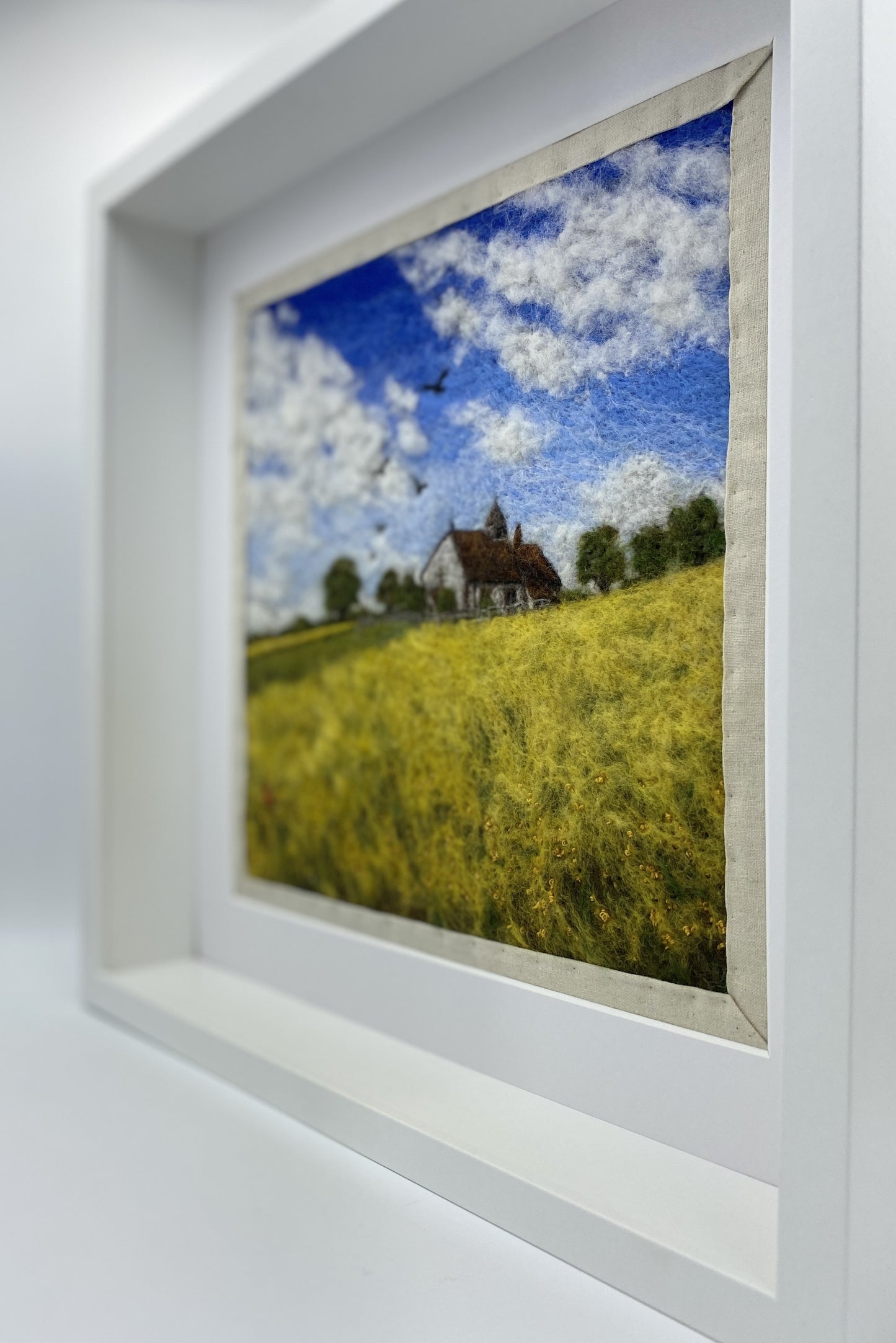 needle felted picture of a Medieval church in a field surrounded by yellow flowers with a blue sky above