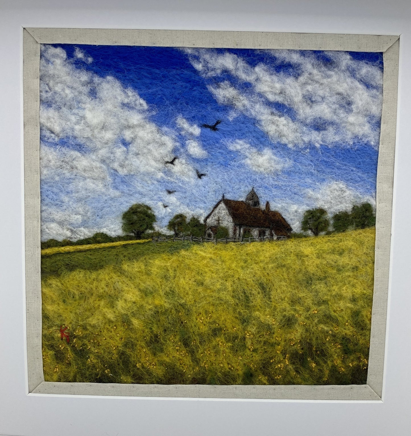 needle felted picture of a Medieval church in a field surrounded by yellow flowers with a blue sky above