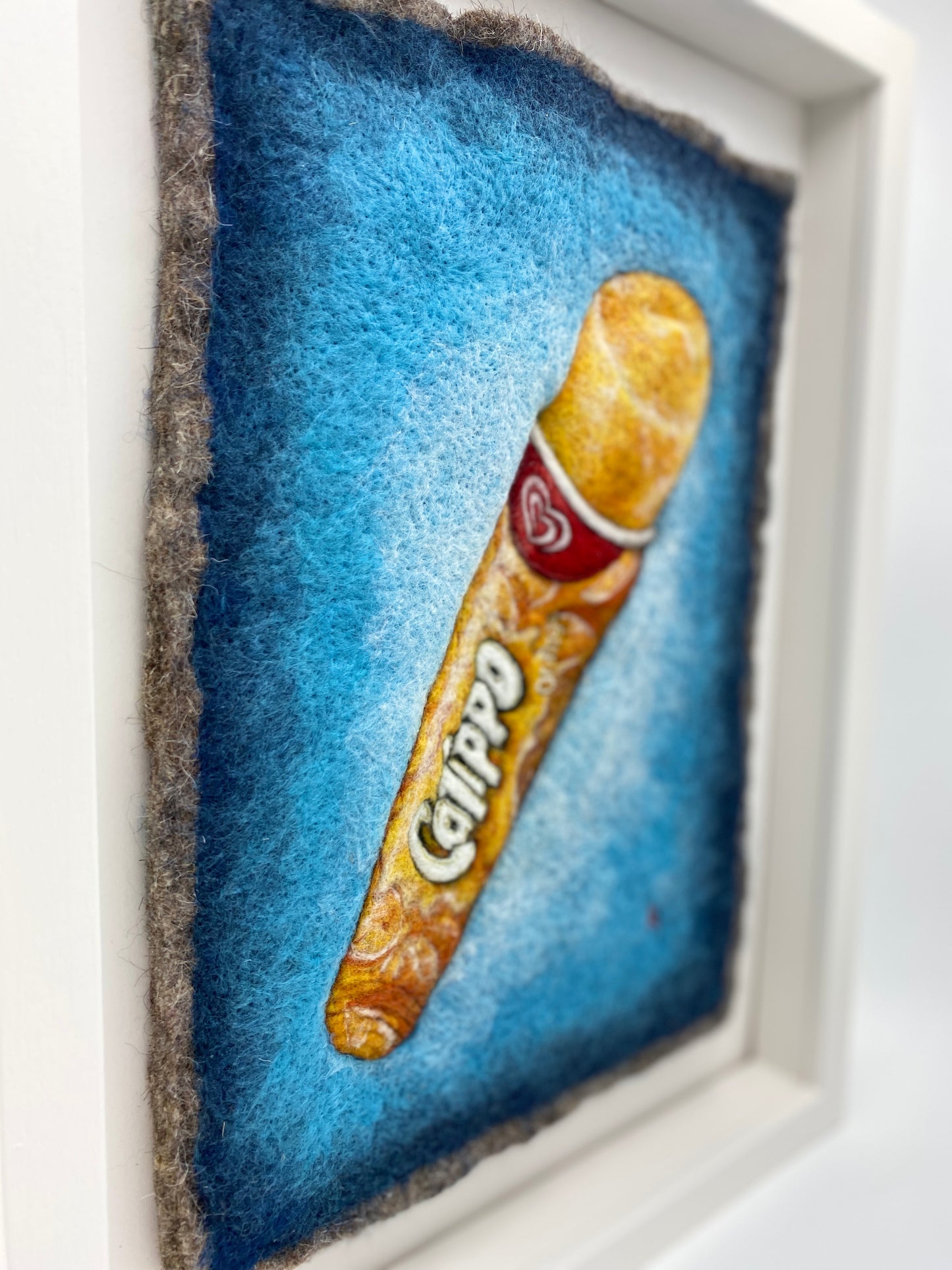 a white box framed needle felted study of an orange flavoured Calippo ice lolly on a blue background