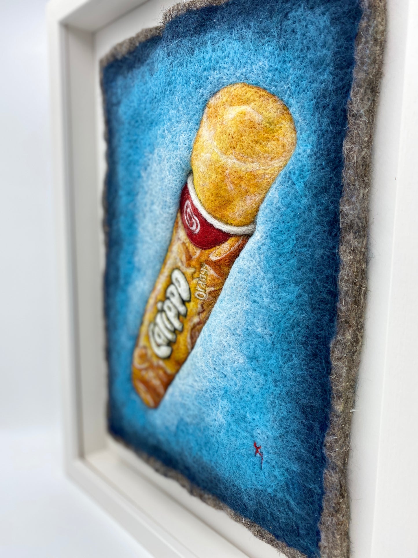 a white box framed needle felted study of an orange flavoured Calippo ice lolly on a blue background
