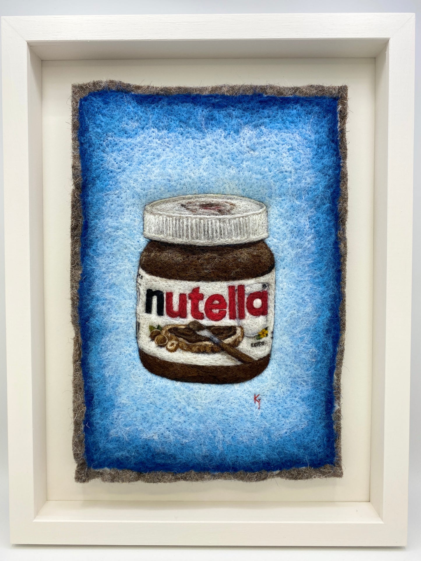 A white wooden framed needle felted picture of a jar of Nutella on a bright blue background