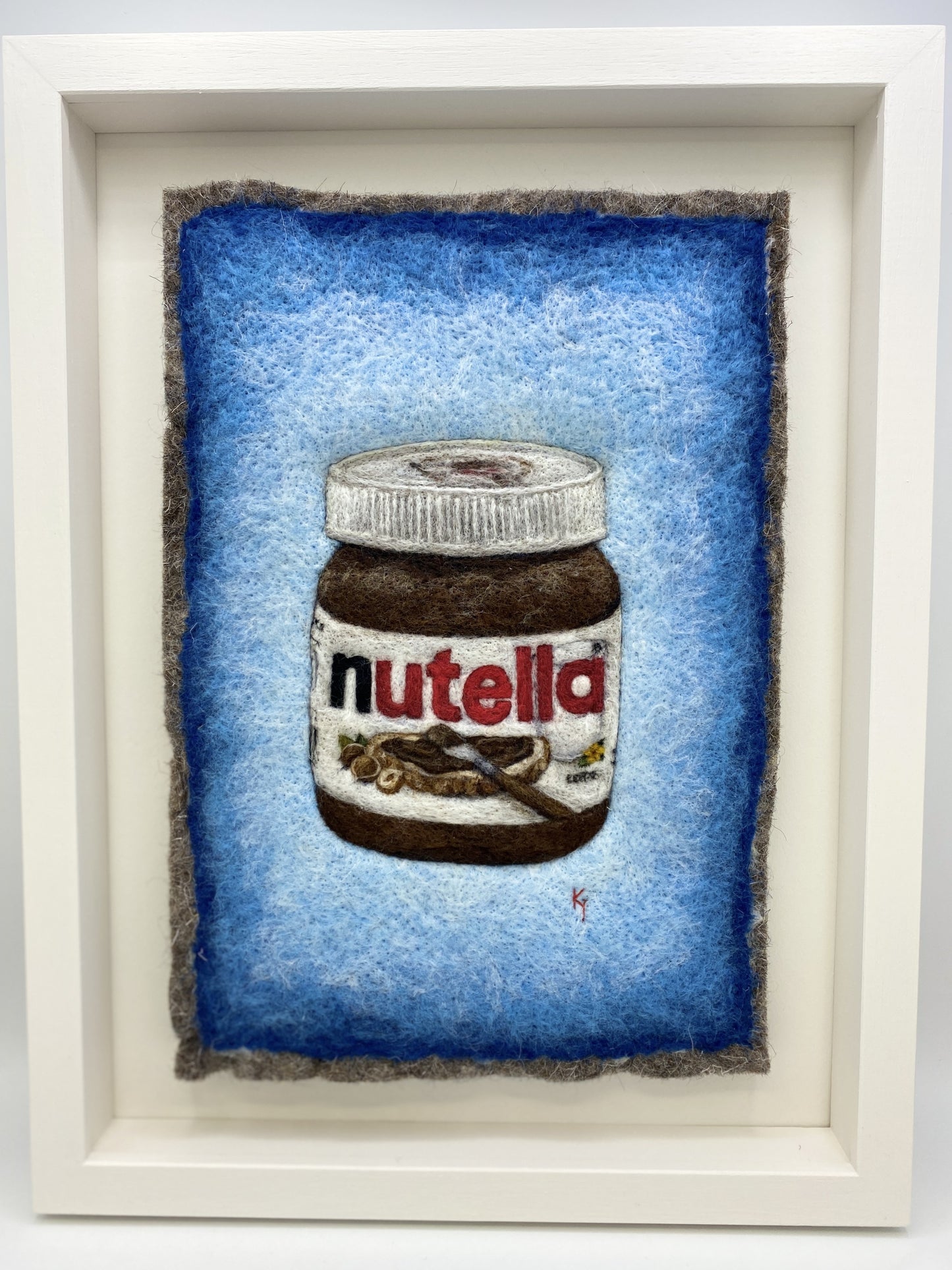 A white wooden framed needle felted picture of a jar of Nutella on a bright blue background