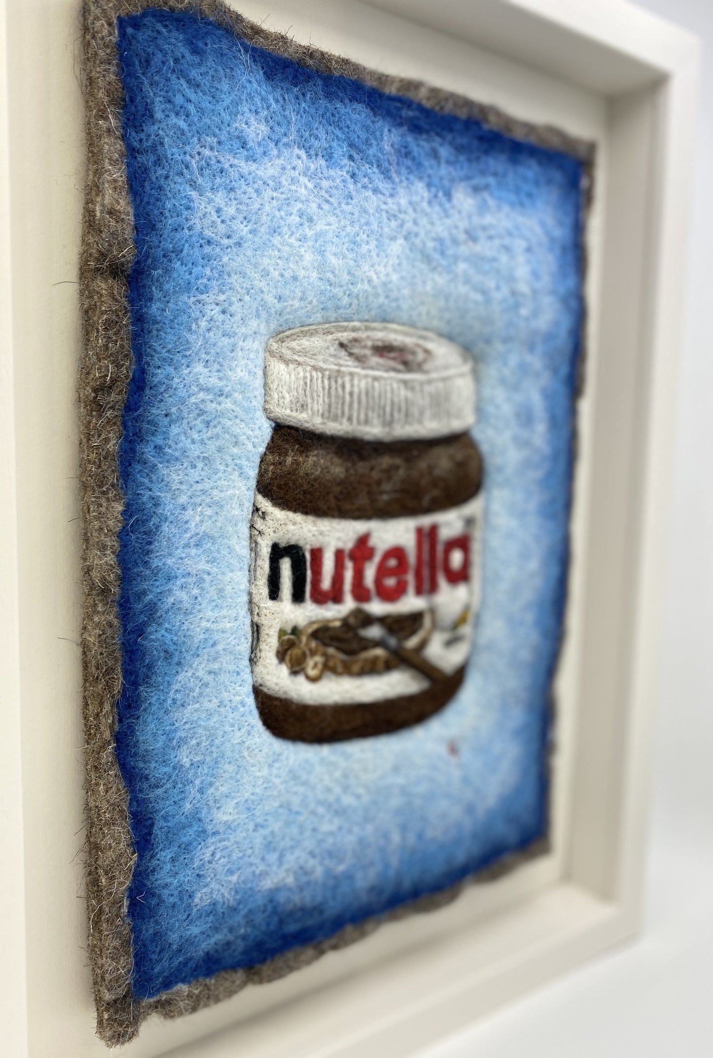 A white wooden framed needle felted picture of a jar of Nutella on a bright blue background