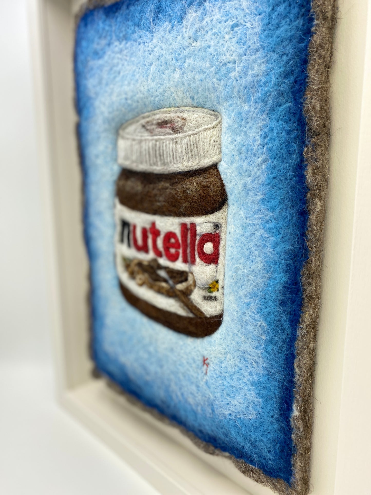 A white wooden framed needle felted picture of a jar of Nutella on a bright blue background