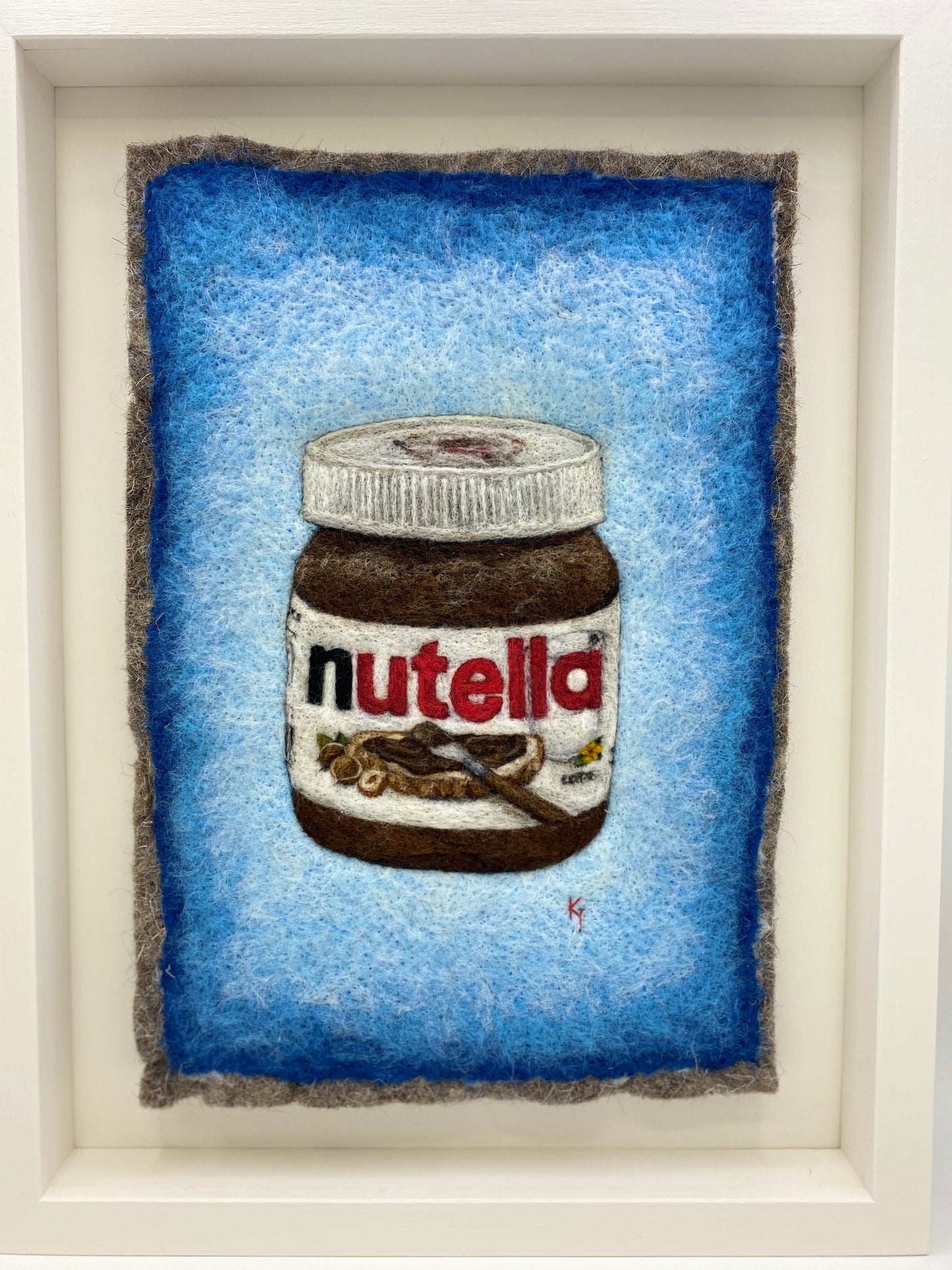 A white wooden framed needle felted picture of a jar of Nutella on a bright blue background