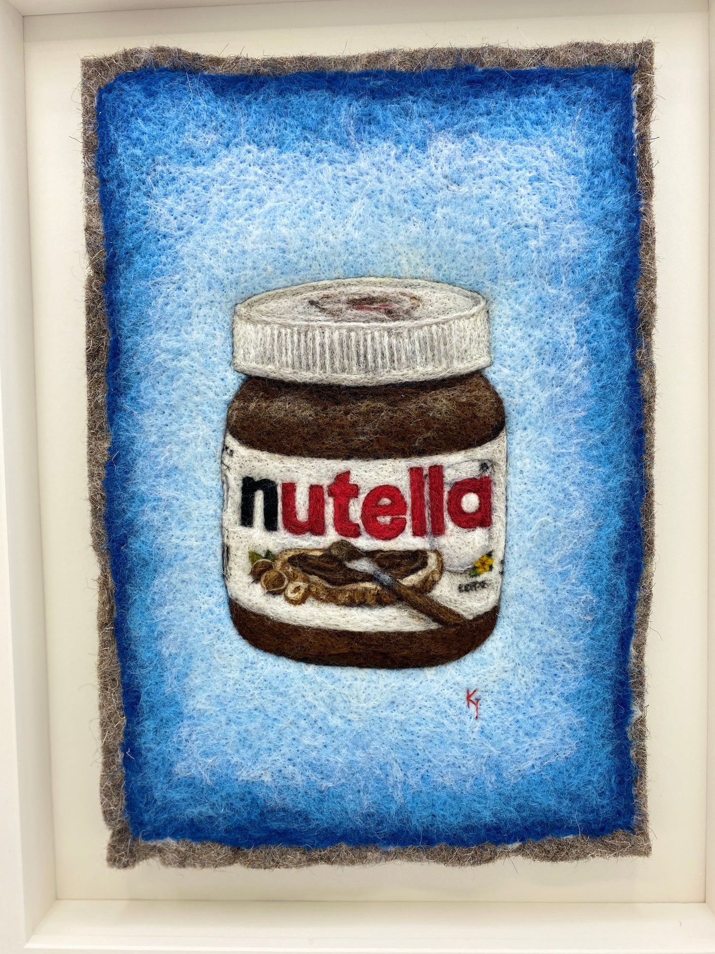 A white wooden framed needle felted picture of a jar of Nutella on a bright blue background