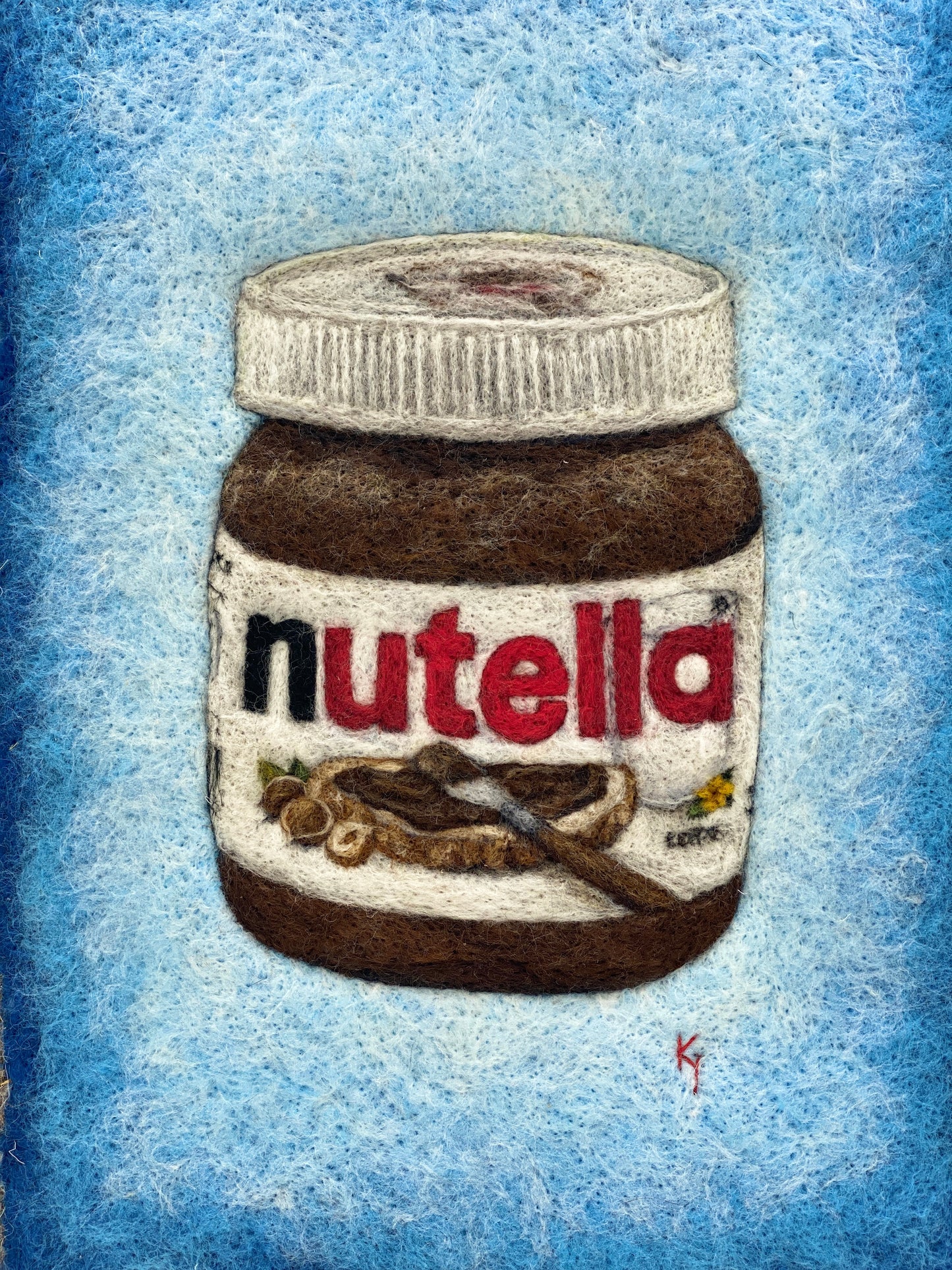 A white wooden framed needle felted picture of a jar of Nutella on a bright blue background