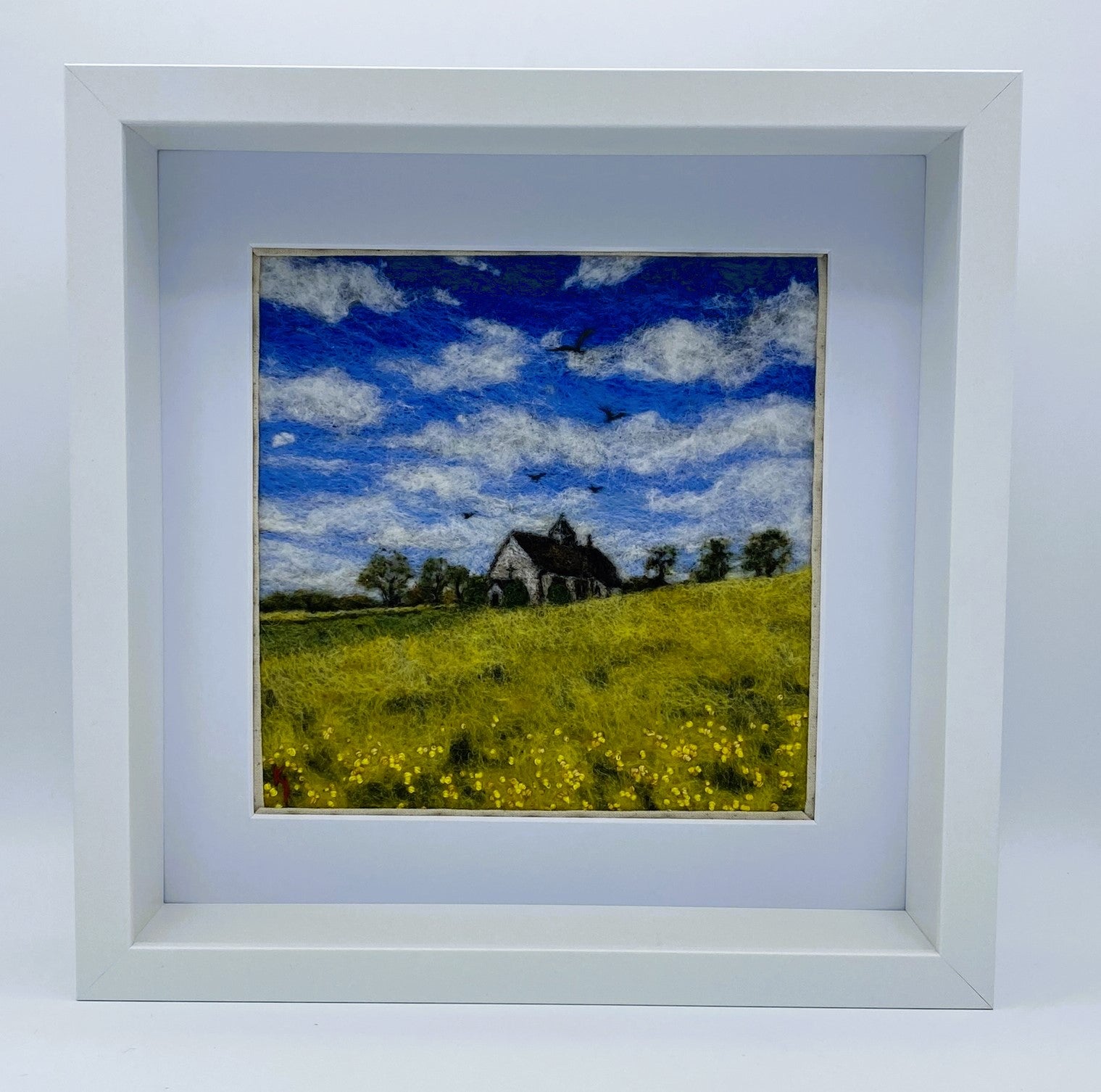 needle felted study of a church (St Hubert's) on a hill surrounded by yellow fields, with a blue cloudy sky and five birds taking flight above the church