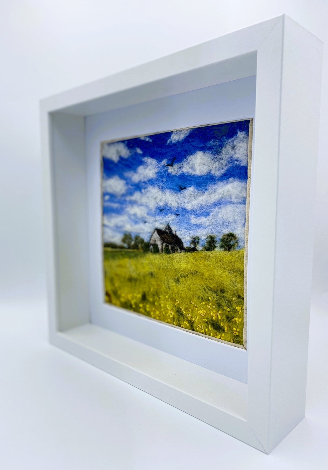 needle felted study of a church (St Hubert's) on a hill surrounded by yellow fields, with a blue cloudy sky and five birds taking flight above the church