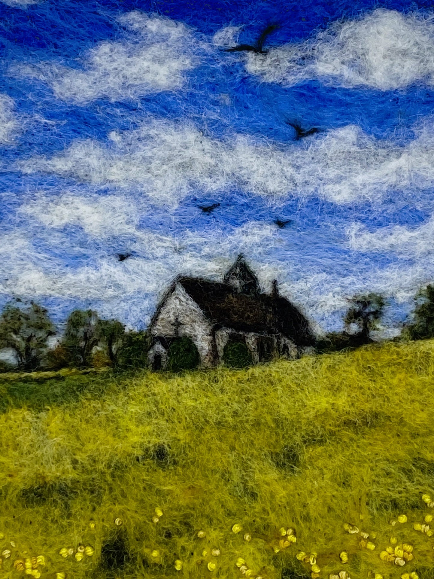 needle felted study of a church (St Hubert's) on a hill surrounded by yellow fields, with a blue cloudy sky and five birds taking flight above the church