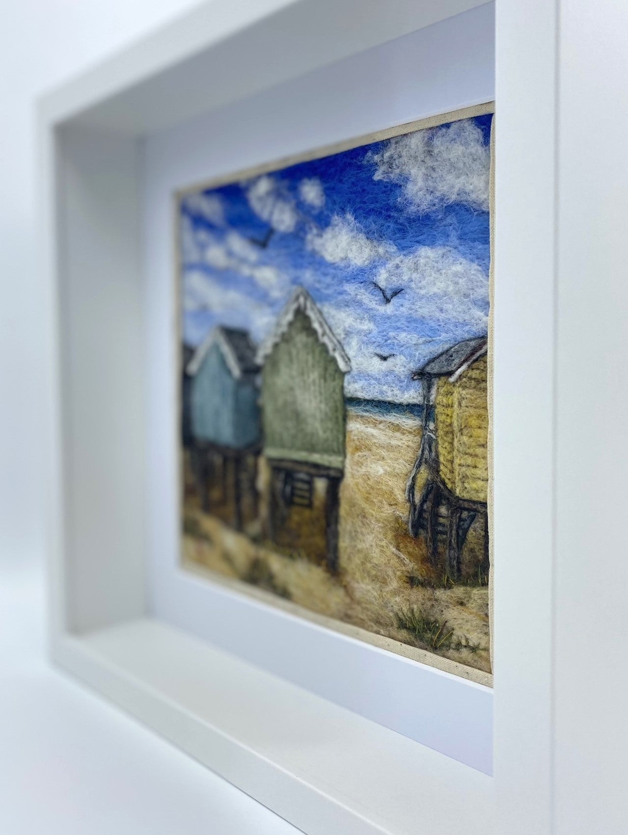 Needle felted picture of four beach huts on stilts facing a sandy beach, blue sea and cloudy blue sky