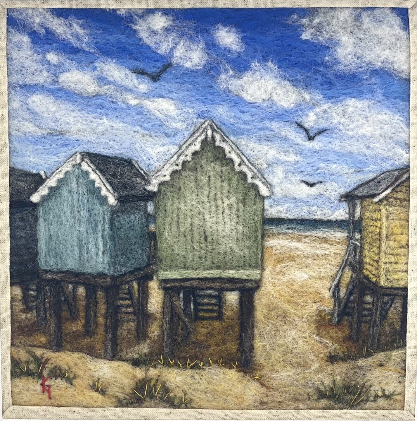 Needle felted picture of four beach huts on stilts facing a sandy beach, blue sea and cloudy blue sky