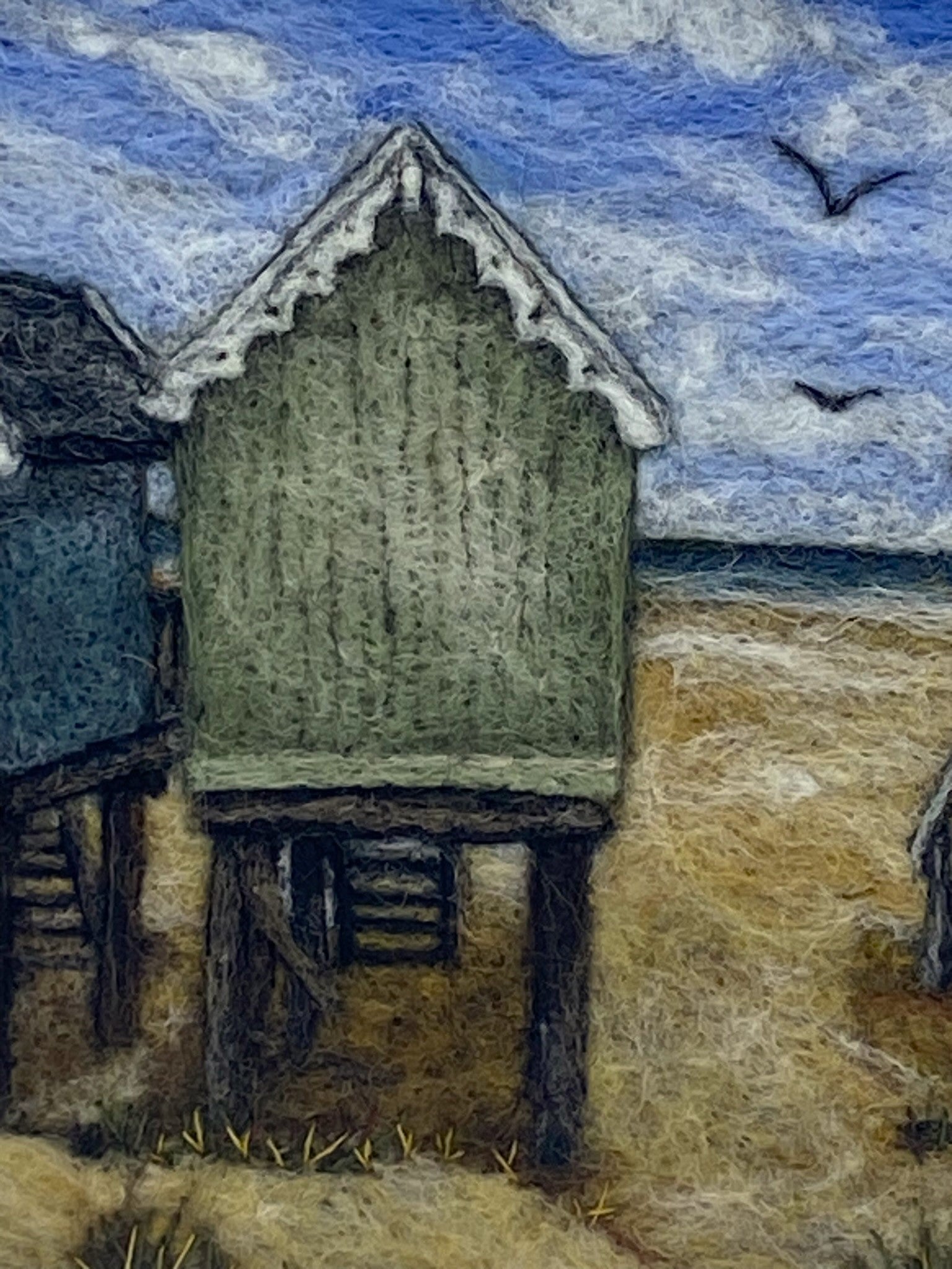 Needle felted picture of four beach huts on stilts facing a sandy beach, blue sea and cloudy blue sky