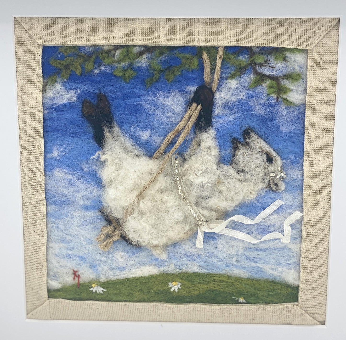 needle felted picture of a sheep on a swing wearing a beaded ribbon belt