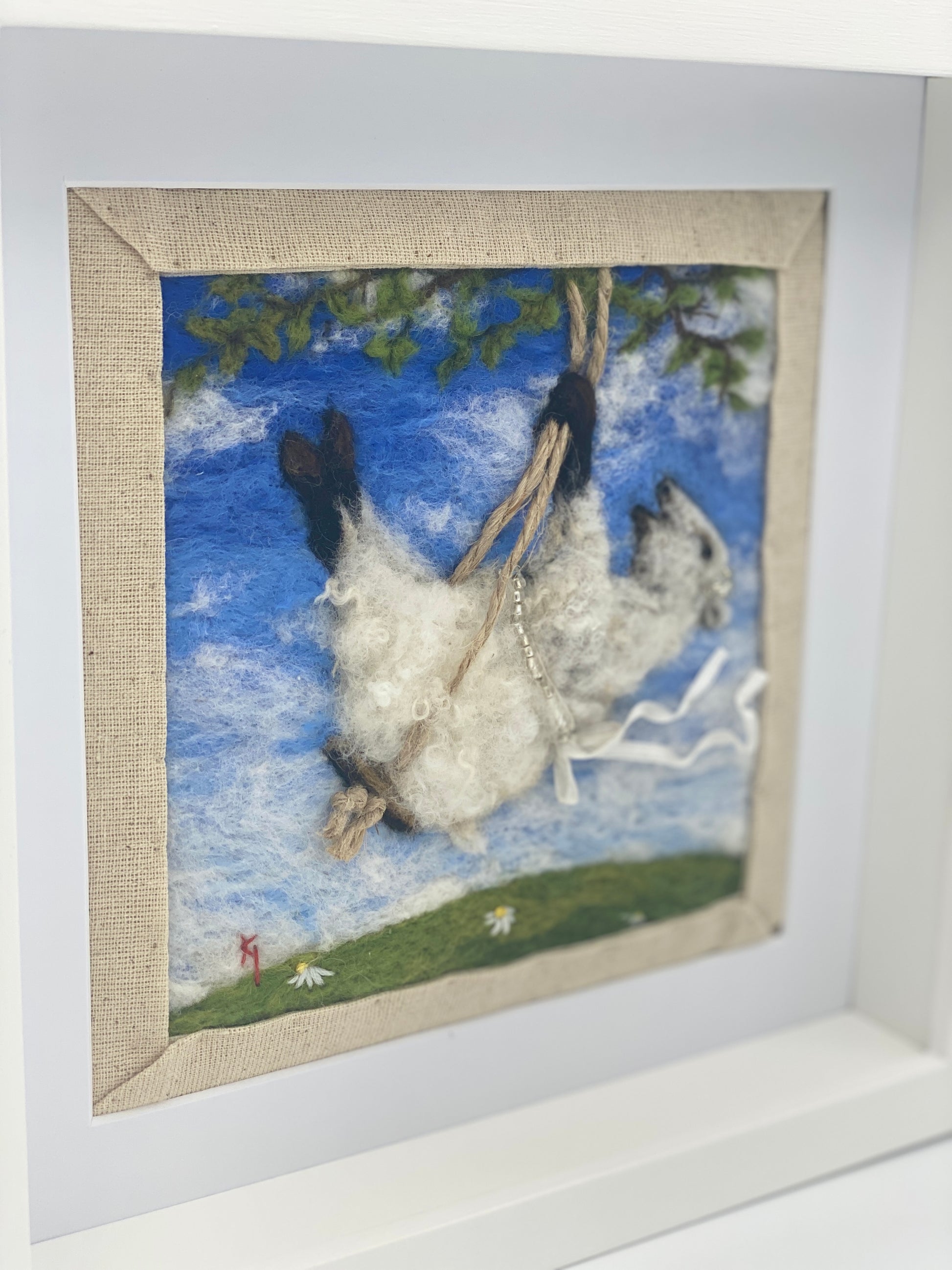 needle felted picture of a sheep on a swing wearing a beaded ribbon belt