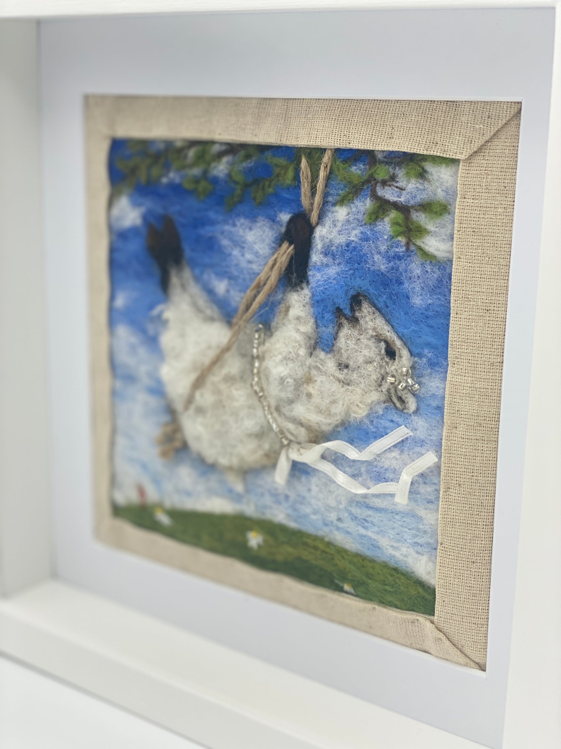 needle felted picture of a sheep on a swing wearing a beaded ribbon belt