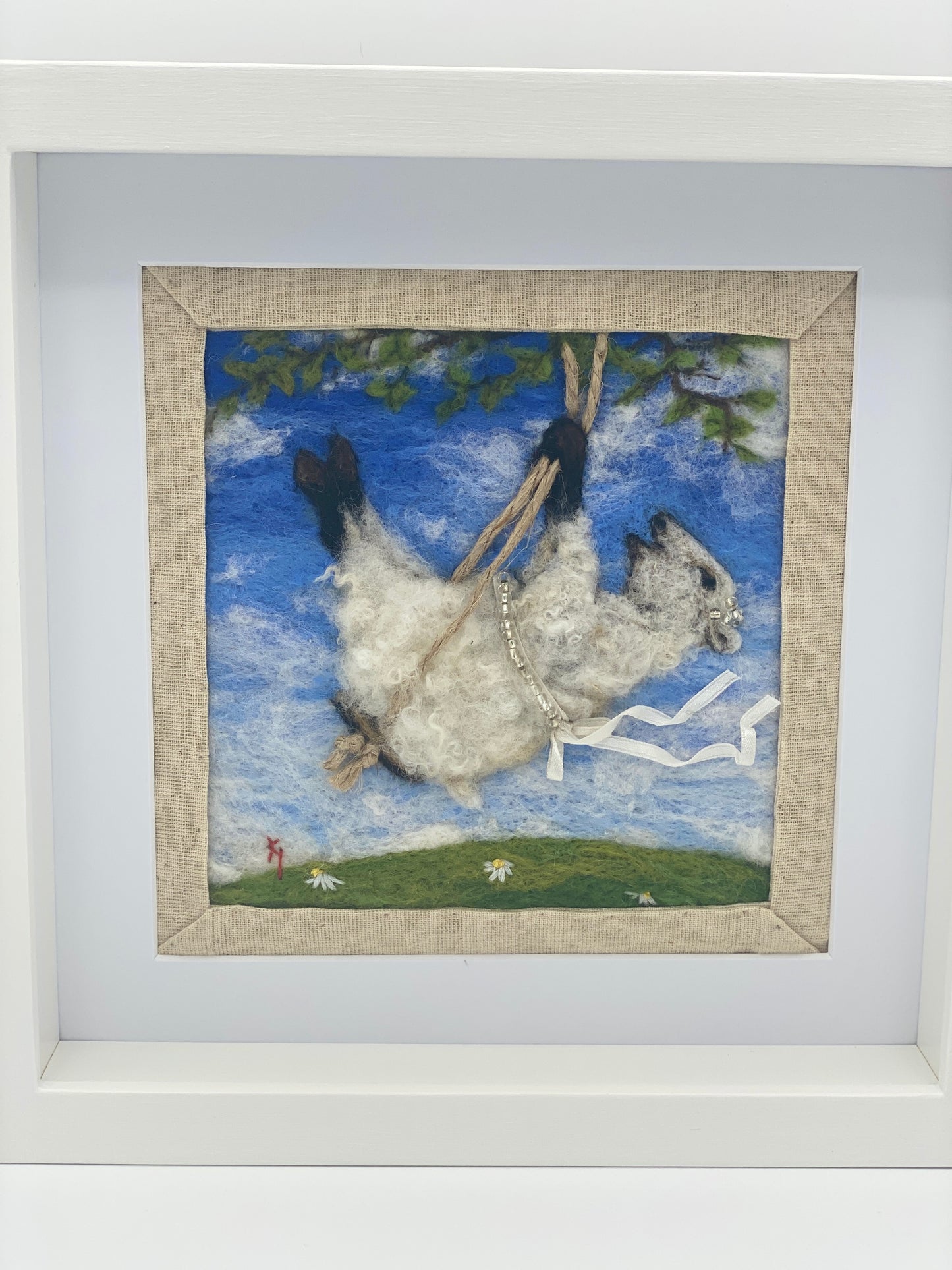 needle felted picture of a sheep on a swing wearing a beaded ribbon belt