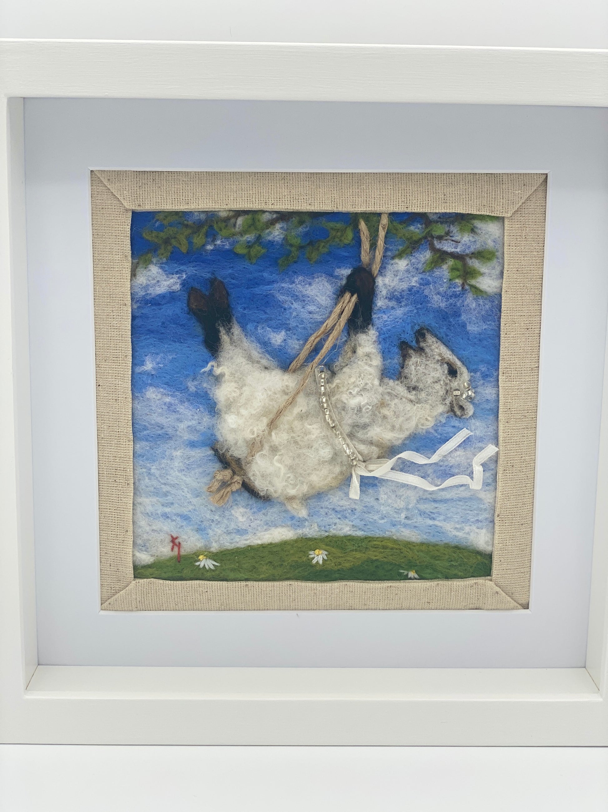 needle felted picture of a sheep on a swing wearing a beaded ribbon belt