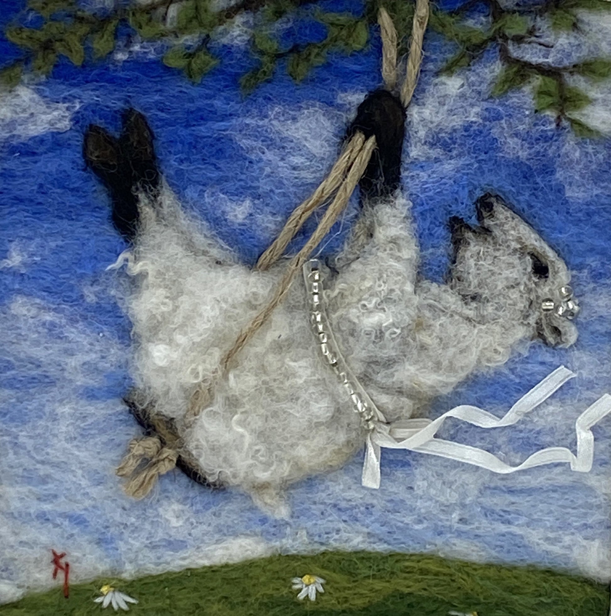 needle felted picture of a sheep on a swing wearing a beaded ribbon belt