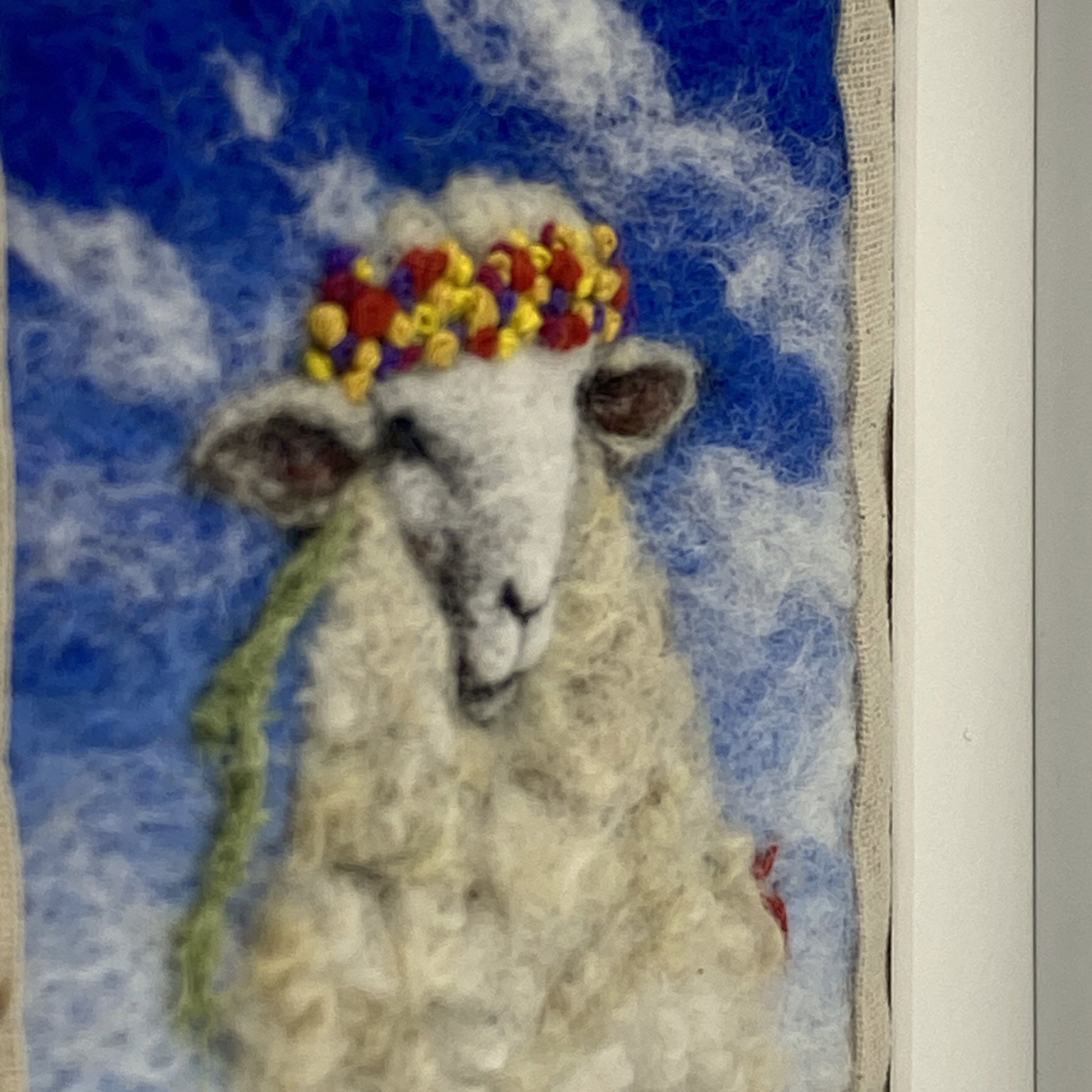 needle felted portrait of a sheep wearing a mulitcoloured stitched crown on a sky blue floral background