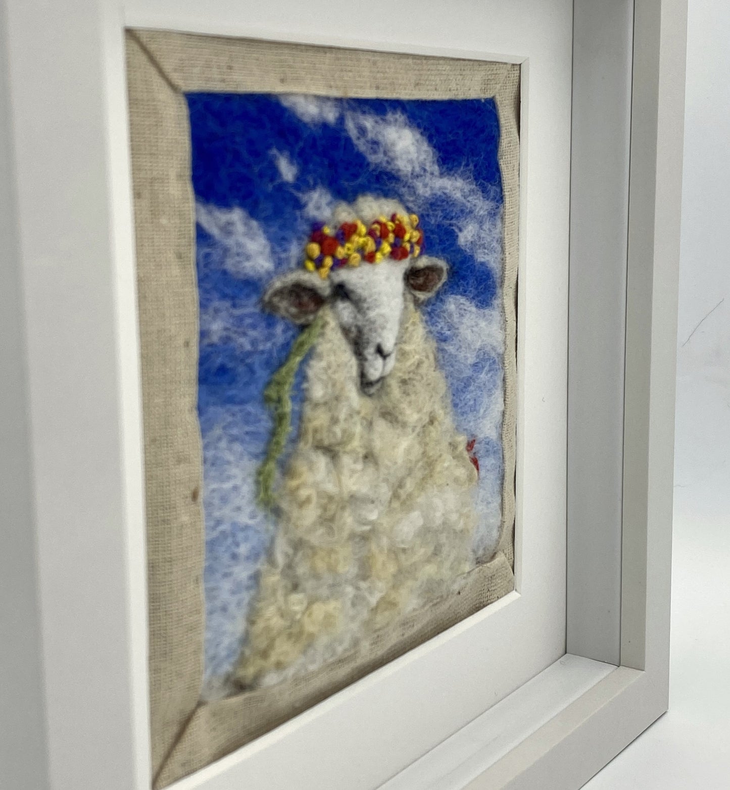 needle felted portrait of a sheep wearing a mulitcoloured stitched crown on a sky blue floral background