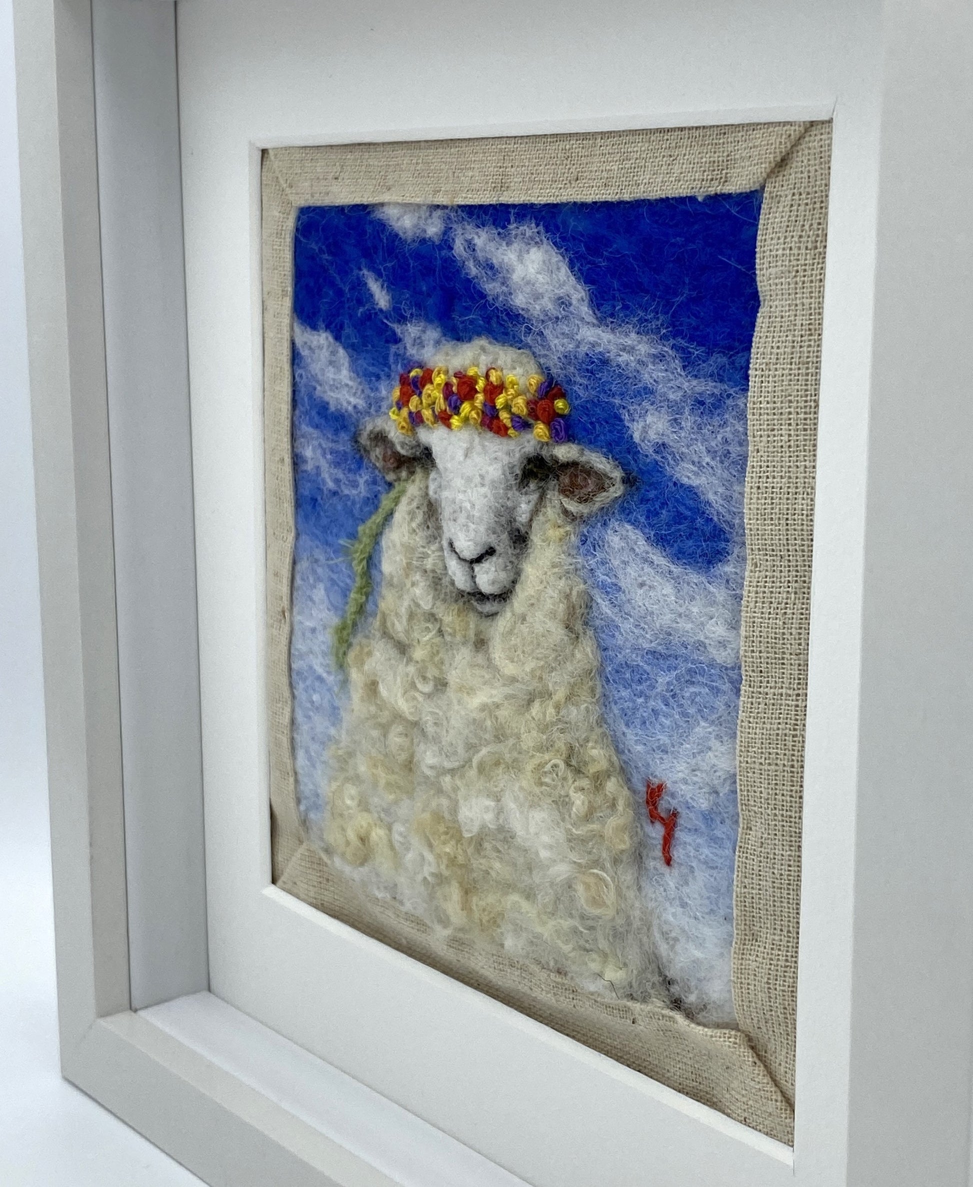 needle felted portrait of a sheep wearing a mulitcoloured stitched crown on a sky blue floral background