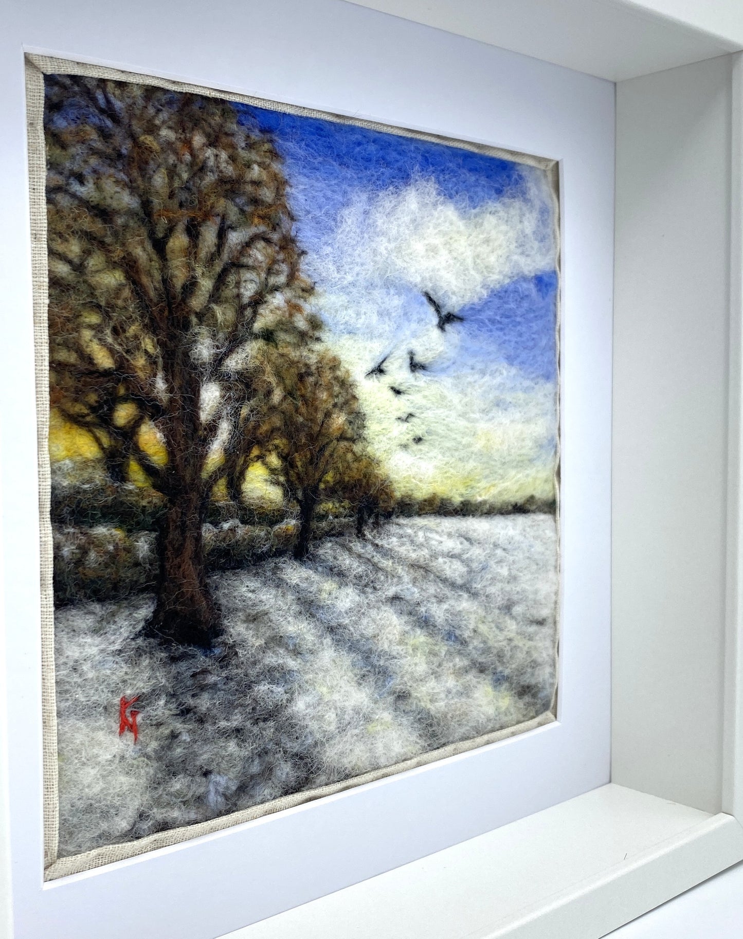 Felted artwork of a landscape with trees and birds in a white frame.