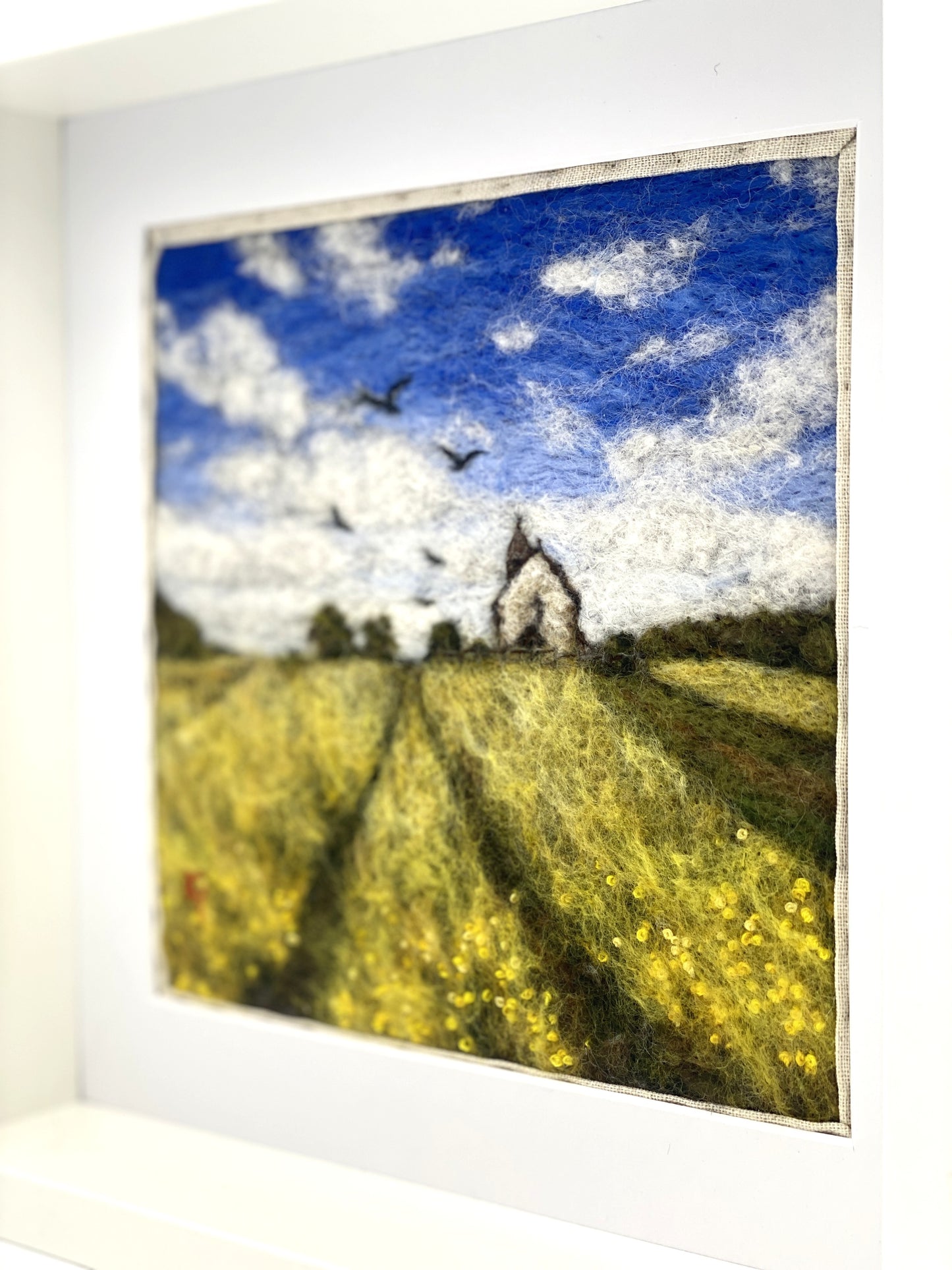 needle felted depiction of a church in a field with birds flying overhead in th cloudy blue sky
