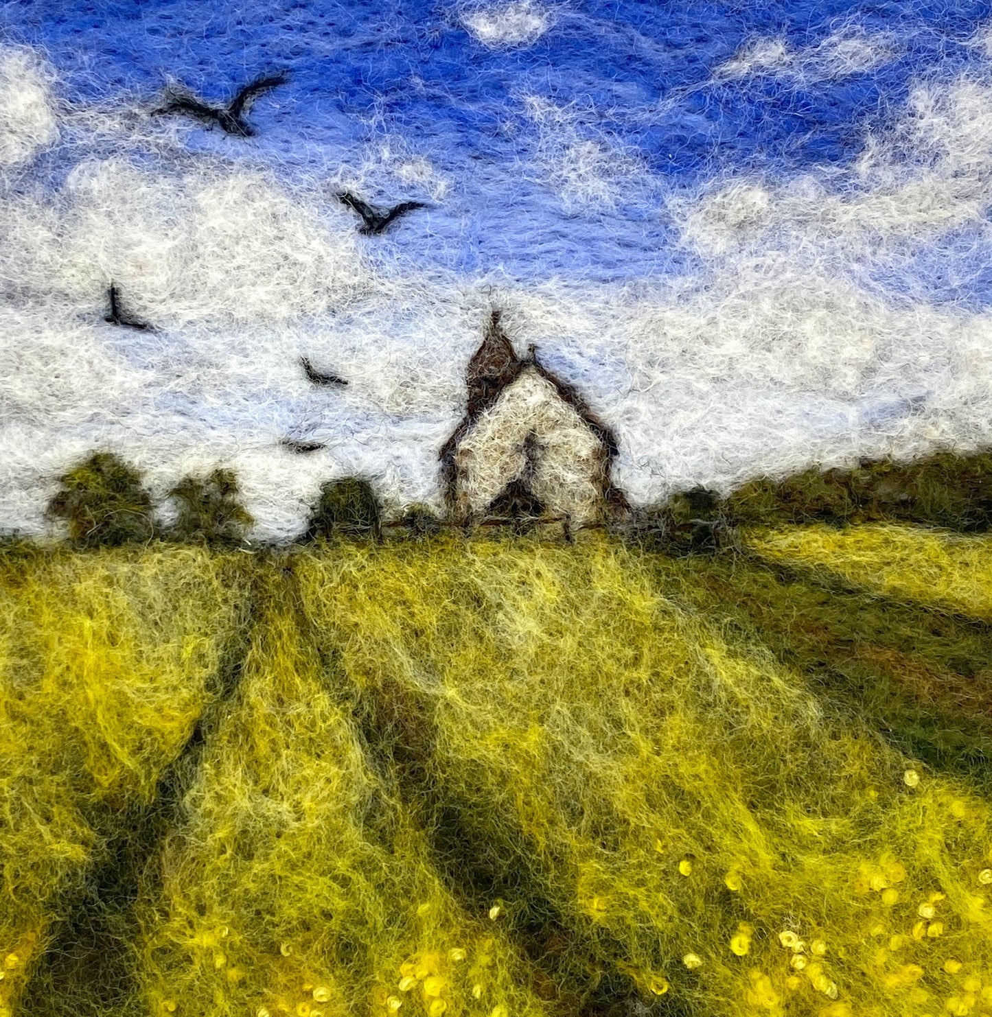 needle felted depiction of a church in a field with birds flying overhead in th cloudy blue sky
