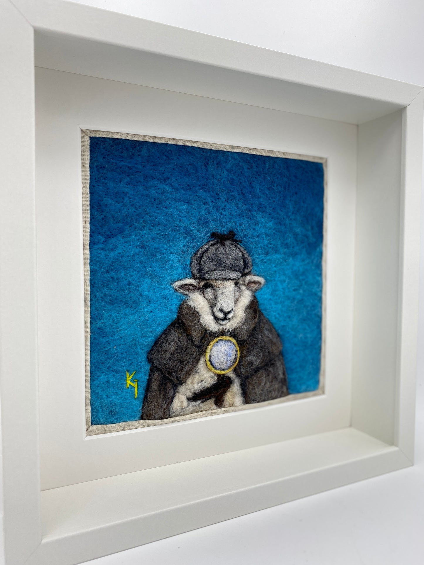 needle felted framed art work of a sheep dressed as Shelock Holmes in coat, deer stalker hat and holding a magnifying glass