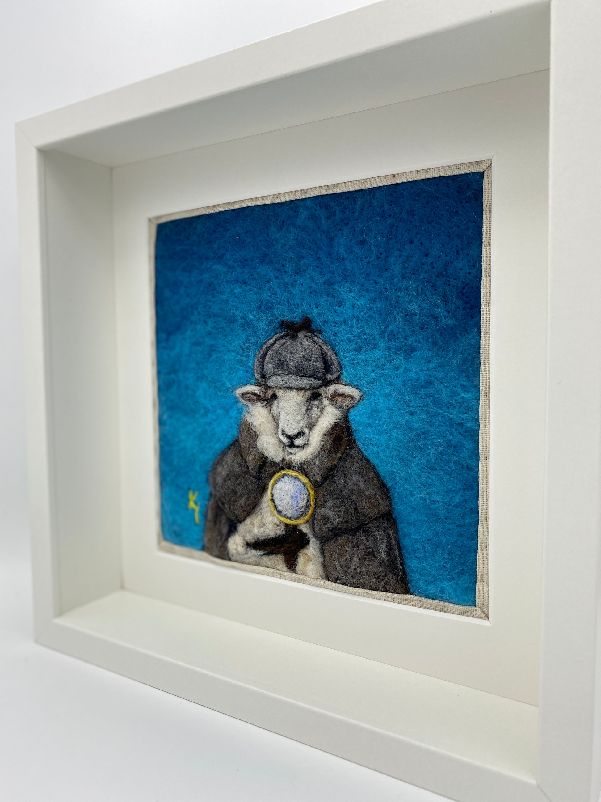 needle felted framed art work of a sheep dressed as Shelock Holmes in coat, deer stalker hat and holding a magnifying glass