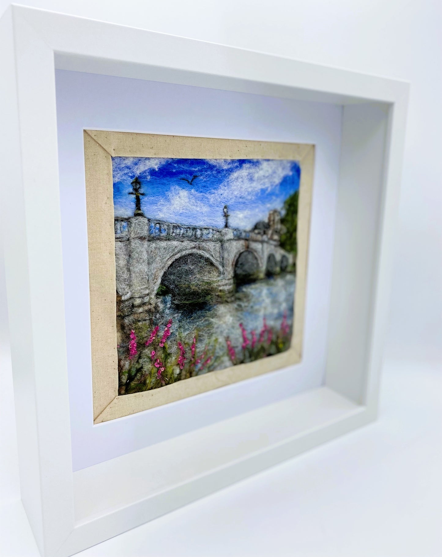 a box-framed, needle felted wool fibre picture of Ricmond Bridge showing the bridge, buildings across the river, trees, lamp posts and embroidered purple wild flowers