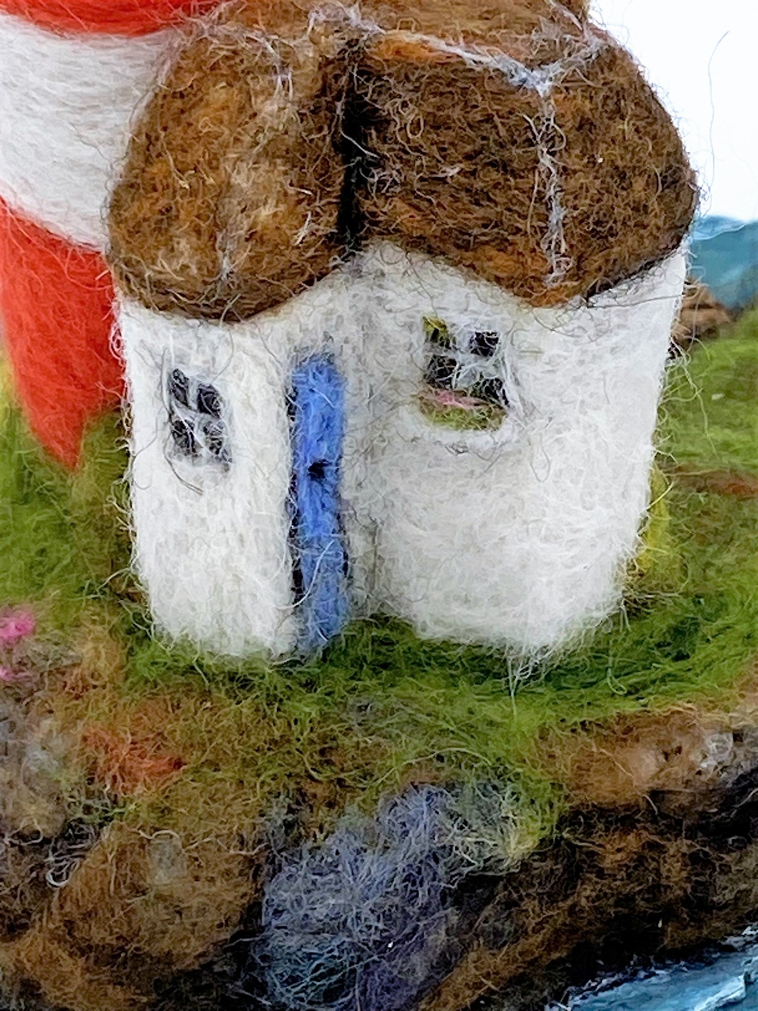 close up of needle felted red and white striped lighthouse on rocky island, sea, beach, white house, washing line, wooden dock and woollen boat